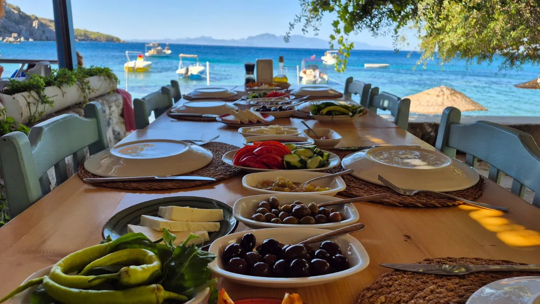 Pruva’nın deniz kıyısındaki uzun sofrasında zeytin, peynir ve mevsim sebzeleriyle hazırlanmış kahvaltı; arkada Mazı koyu ve tekneler